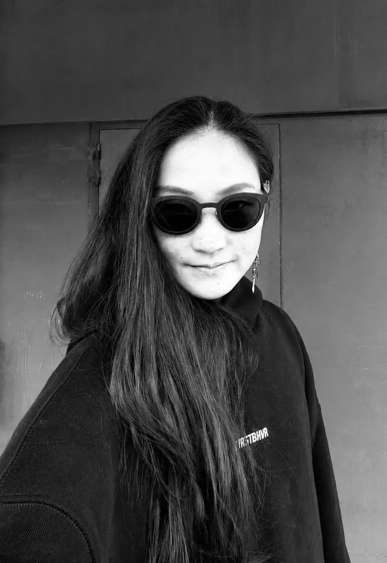 Black-and-white portrait of Jasmine Fan in round dark sunglasses and a black sweatshirt, her long dark hair over one shoulder, in front of a metal door.